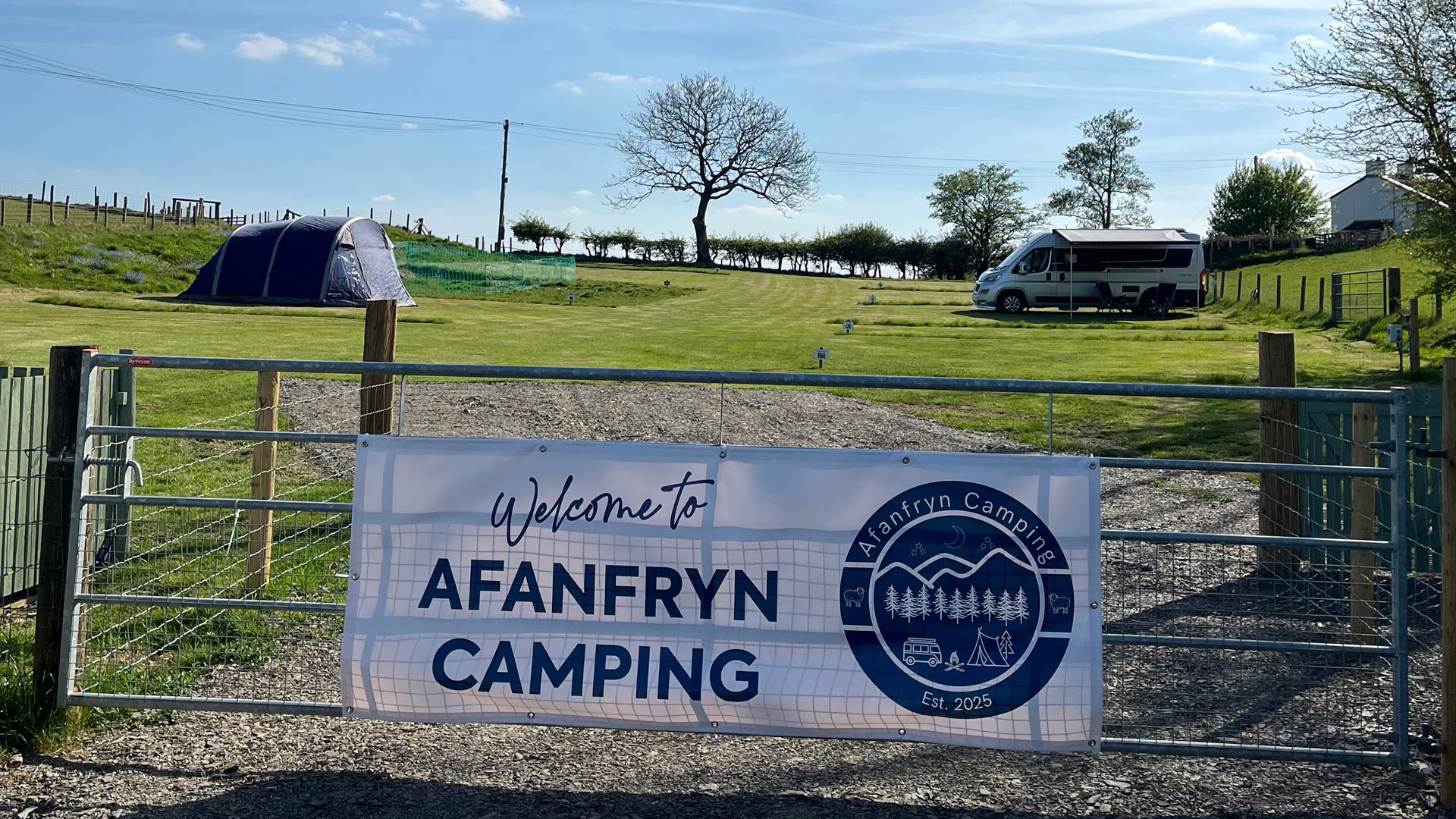 Entrance to Afanfryn Camping. There is an advertising banner on the metal gate with a freshly-mown grassed area containing a tent and a camper van with awning. There are trees and hedge in the background